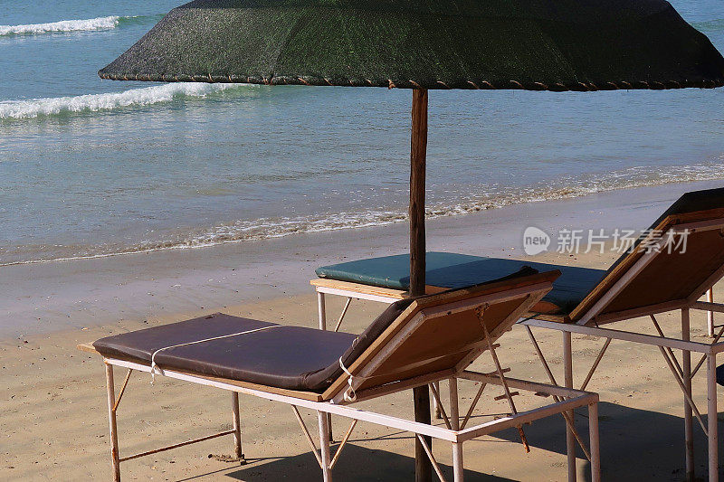 在热带沙滩上，木制太阳躺椅撑着阳伞，沙滩浴巾晒日光浴，在树荫下睡觉，木制太阳躺椅热带海滩假日海滨度假印度果阿/喀拉拉邦